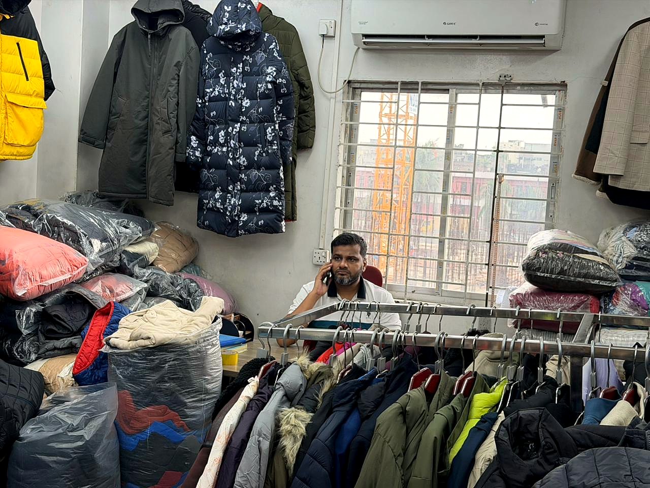 A man on a phone call at a desk in the Sakib office, behind a rail of coats, with bagged stock and hanging jackets around him